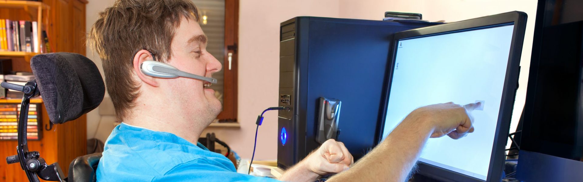 Man with infantile cerebral palsy using a computer with a wireless headset, reaching out to touch the touch screen